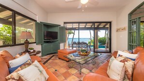 A living room with a TV, green entertainment unit, orange sofa, and a glass table. - Beautiful Hacienda style villa located on white sandy beach, pool, AC, WiFi! (South Akumal)