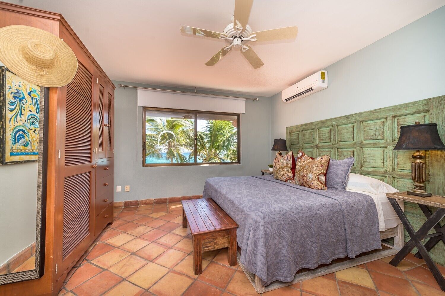 A bedroom with a bed, a window with a view of palm trees, a ceiling fan, and a wooden headboard.