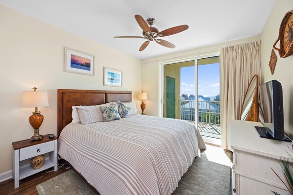 Une chambre à coucher avec un grand lit, deux lampes de chevet, un ventilateur de plafond, une télévision et une vue sur l’extérieur.