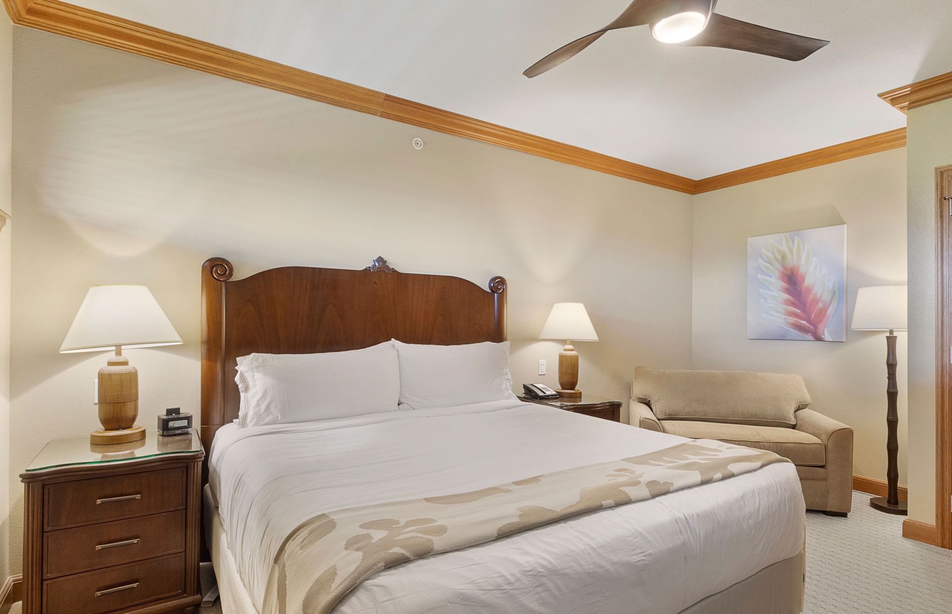 Une chambre d’hôtel avec un lit doté d’une tête de lit en bois, deux lampes de chevet, un fauteuil beige et un ventilateur de plafond.