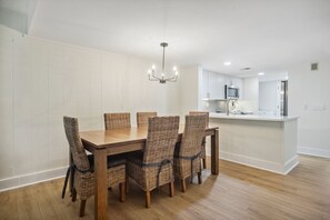 Ein Essbereich mit Holztisch und Korbstühlen, eine Küche mit weißen Schränken und einer Mikrowelle sowie eine Deckenleuchte.