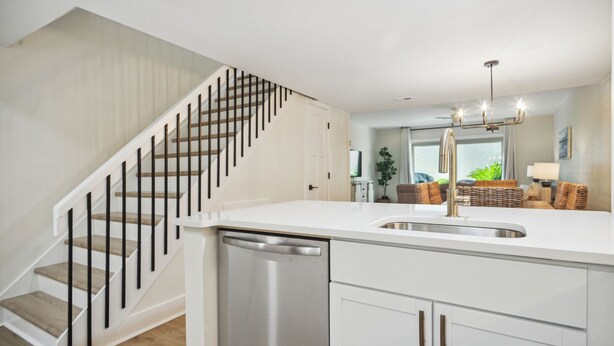 A modern kitchen with a white countertop, stainless steel sink, and a dishwasher. There is a staircase with wooden steps and black railings leading to an upper level. - Surf Court 60 - 3 Story South Forest Beach Townhouse (Hilton Head Island)