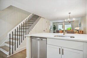 A modern kitchen with a white countertop, stainless steel sink, and a dishwasher. There is a staircase with wooden steps and black railings leading to an upper level. - Surf Court 60 - 3 Story South Forest Beach Townhouse (Hilton Head Island)