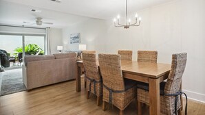 A dining area with a wooden table and wicker chairs, a ceiling fan, and a view of the outdoors through a sliding door. - Surf Court 60 - 3 Story South Forest Beach Townhouse (Hilton Head Island)