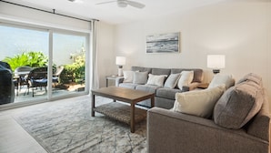 A modern living room with a sofa, armchairs, a coffee table, and a ceiling fan. There is a large window with a view of greenery and a patio area with outdoor furniture. - Surf Court 60 - 3 Story South Forest Beach Townhouse (Hilton Head Island)