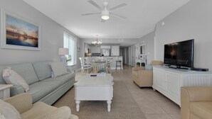 A spacious living room with a sofa, a coffee table, and a television. There is a dining area with a table and chairs, and a kitchen in the background.