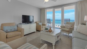 A spacious living room with a flat-screen TV, a ceiling fan, and a large window offering a view of the ocean.
