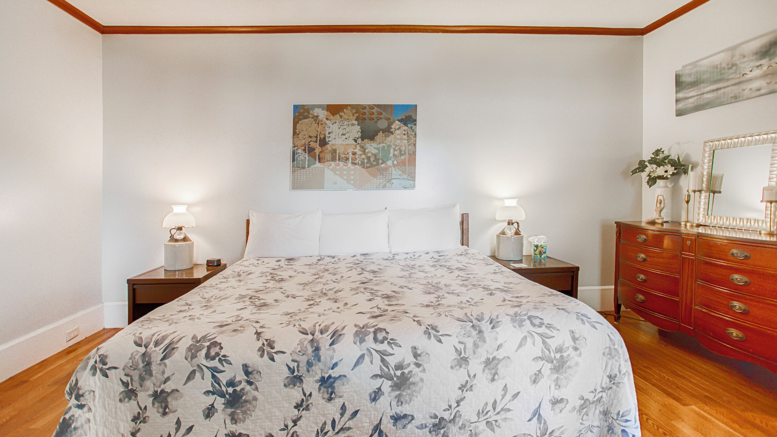 Een slaapkamer met een bloemenbedekking, twee bedlampen, een nachtkastje, een dressoir met laden, en een schilderij aan de muur.