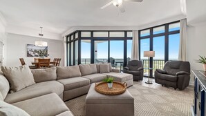 A modern living room with a sectional sofa, a small ottoman, and a dining area with a table and chairs. - The Oasis at Orange Beach 2114 | Lazy River, Water Slide | My Beach Getaways (Orange Beach)