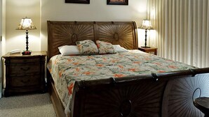 Quarto com cama de madeira, duas mesas de cabeceira com abajures, um criado-mudo, uma janela com cortinas e duas fotos emolduradas na parede.