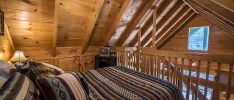 Un dormitorio acogedor en el ático con techo inclinado, cama con ropa de cama estampada, televisor sobre una mesita y una ventana que deja entrar luz natural.