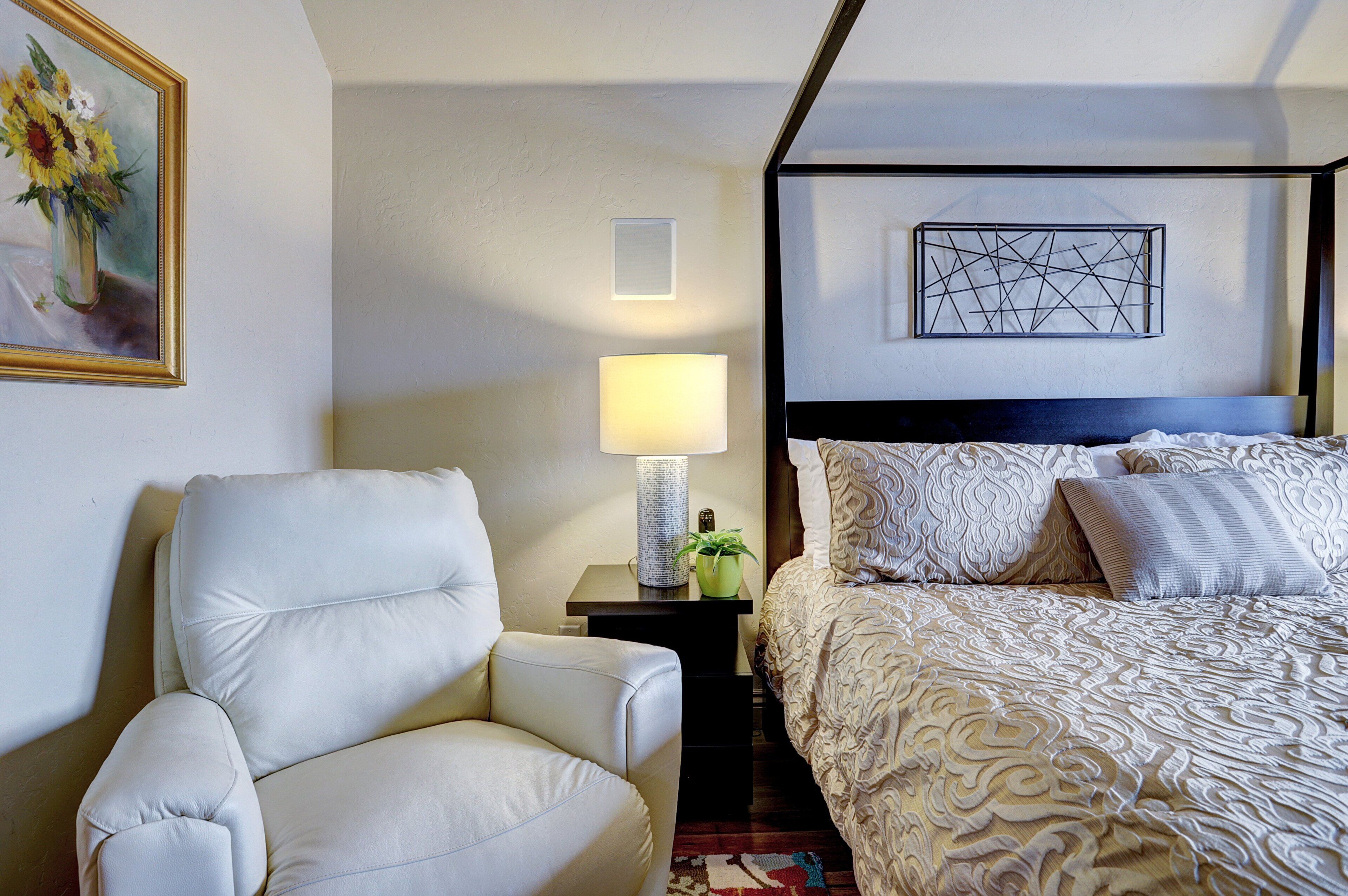 A hotel room with a bed, a chair, a nightstand with a lamp, a vase with flowers, and a painting on the wall.