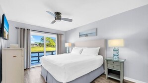 A hotel room with a large bed, a ceiling fan, a nightstand with a lamp, and a view of a body of water through the window. - Bay View, Six-Seat Golf Cart, Pet Friendly, Linkside Village 434 (Miramar Beach)