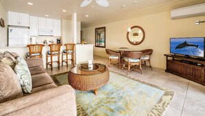 A living room with a sofa, wicker chairs, a coffee table, a TV, and a kitchen in the background.