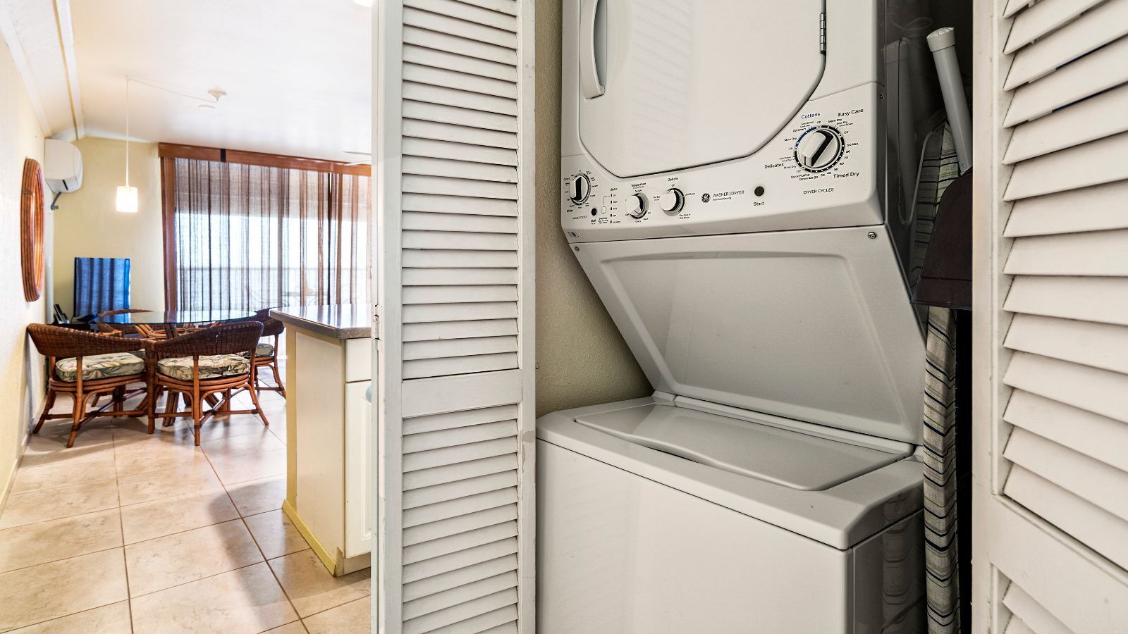 Une buanderie équipée d’un lave-linge et d  ’un sèche-linge, un coin repas avec des chaises en osier et une fenêtre avec des stores.