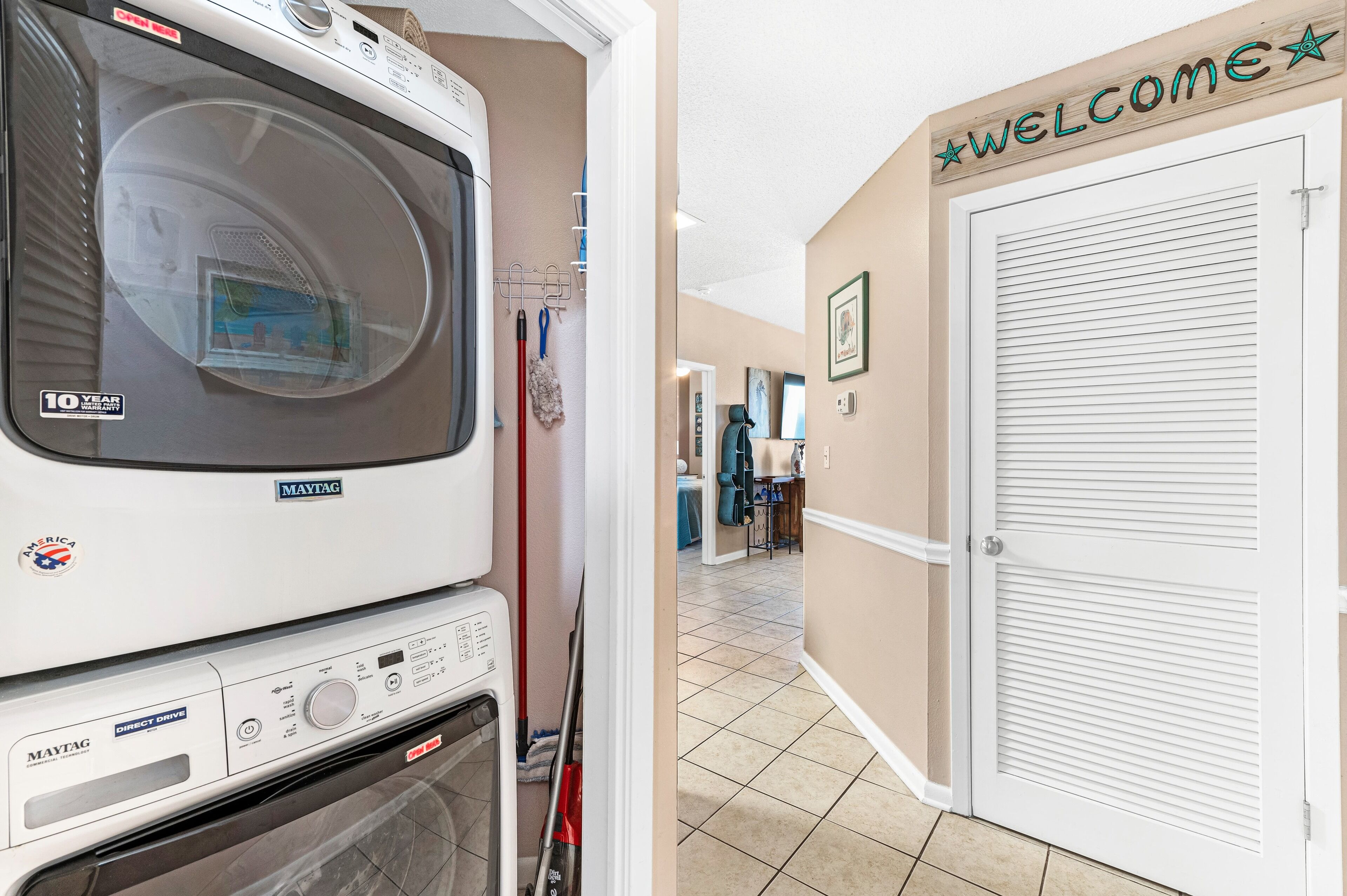 A laundry area with a Maytag washer and dryer, a mop, and a broom. There is a 'WELCOME' sign on the wall and a door with a window.