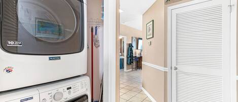 A laundry area with a Maytag washer and dryer, a mop, and a broom. There is a 'WELCOME' sign on the wall and a door with a window.