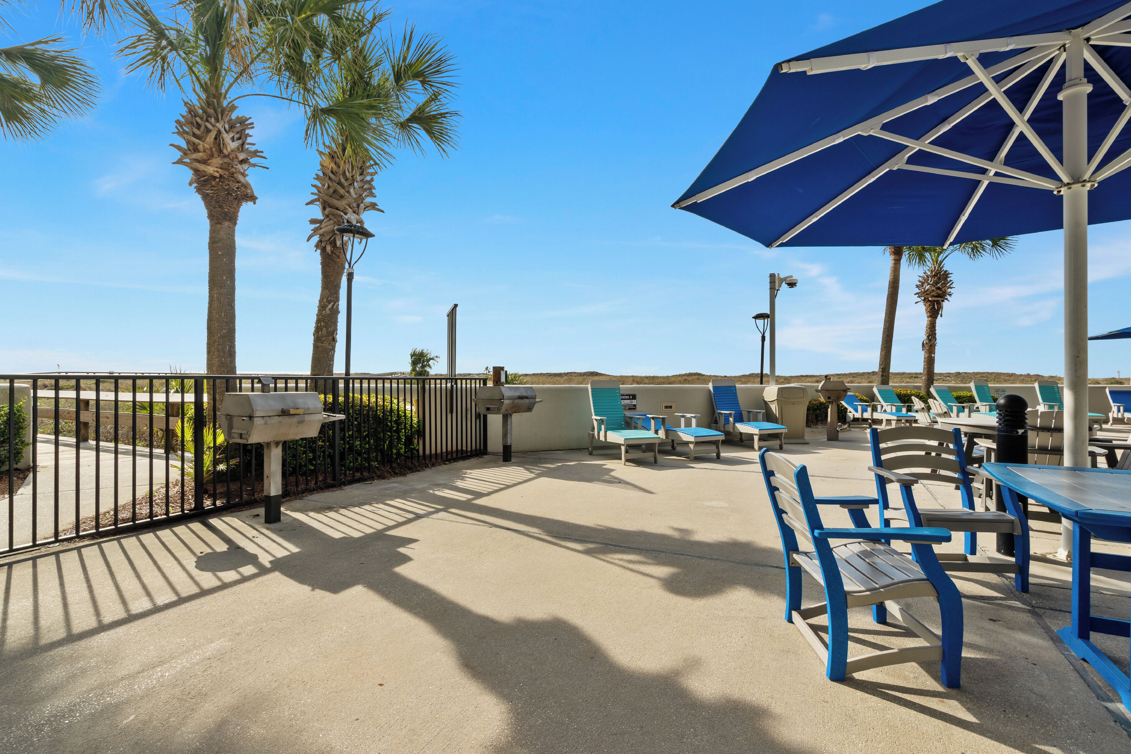 A poolside area with blue and white lounge chairs, a blue umbrella, and palm trees.