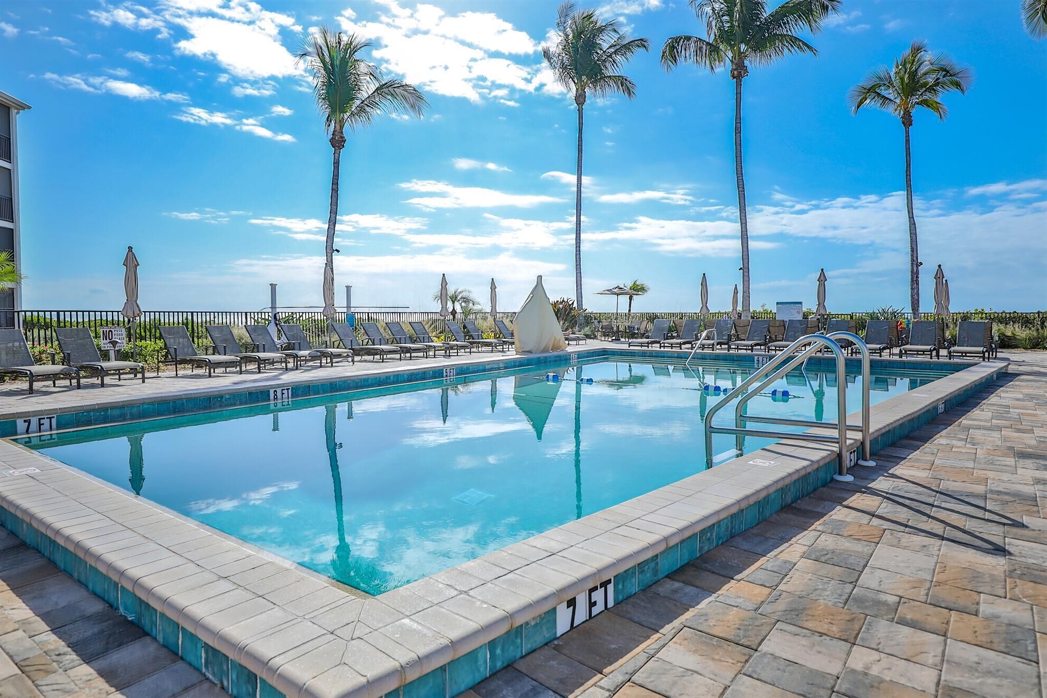 Piscina com espreguiçadeiras e palmeiras, cercada por um deck revestido de azulejos.