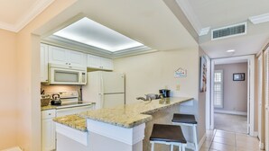 A kitchen with white cabinets, a granite countertop, a microwave, a refrigerator, and a built-in oven. There are two bar stools and a window with blinds. - SUNDIAL D306 - Lovely Sundial Sanibel Condo. Short Walk to Beach (Sanibel)