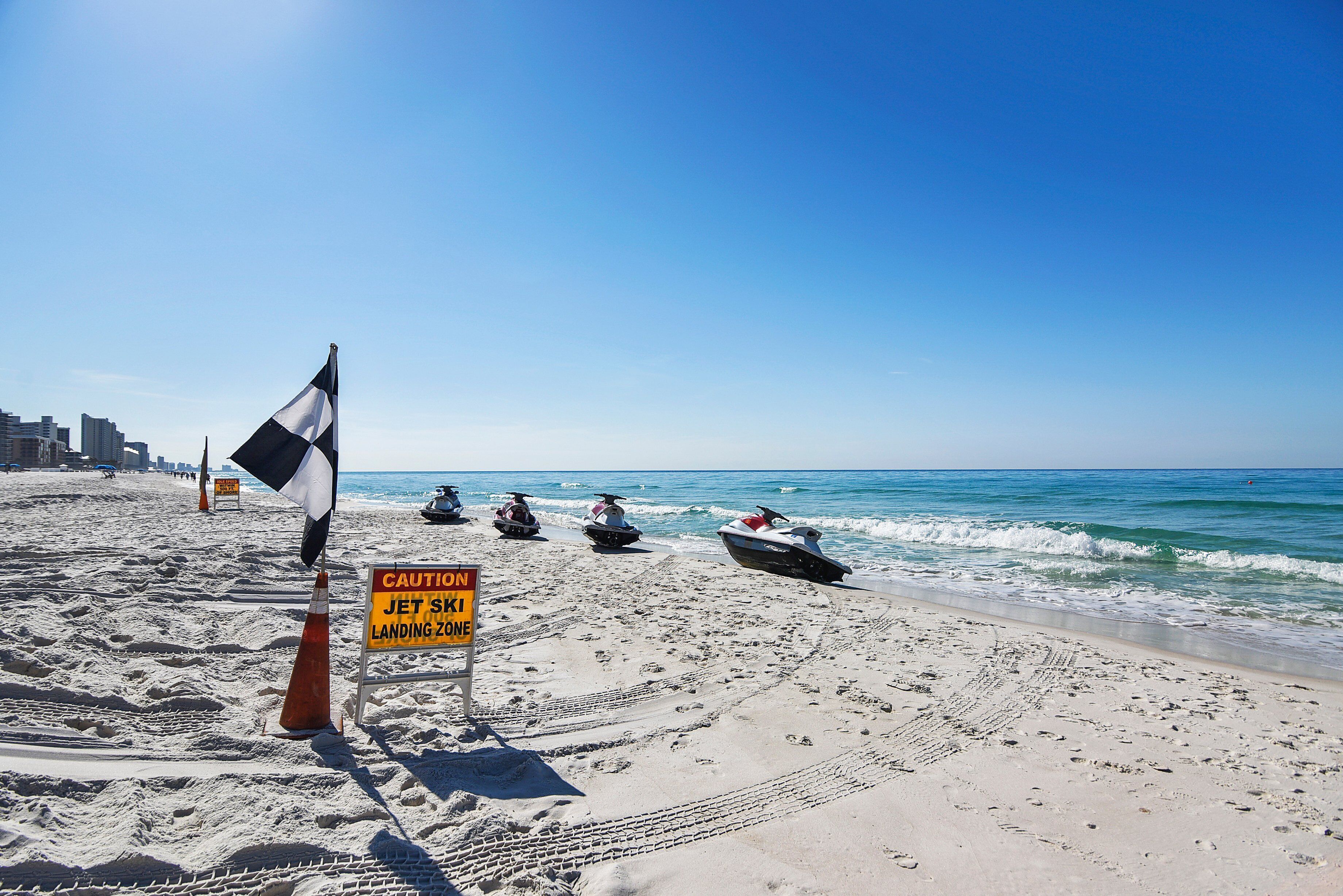 Una playa con motos acuáticas, un letrero de precaución y un cielo azul despejado.