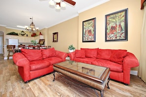Un salon avec un canapé rouge, une table basse en verre, un parquet en bois, un lustre et deux tableaux tropicaux encadrés accrochés au mur.
