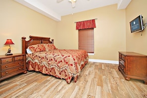 Une chambre à coucher avec un lit en bois, une table de chevet, une commode, une fenêtre avec des stores et un téléviseur fixé au mur.