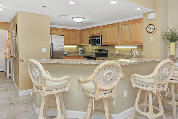 A kitchen with wooden cabinets, a stainless steel refrigerator, a microwave, a stove, and a sink. There are three wicker chairs at the island.