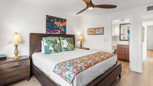 A bedroom with a bed, two bedside tables with lamps, a ceiling fan, a painting, and a bathroom visible through an open door.