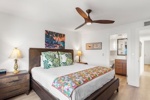 Un dormitorio con cama, dos mesitas de noche con lámparas, un ventilador de techo, un cuadro y un baño visible a través de una puerta abierta.