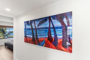 A bedroom with a large painting of a beach scene, a bed, and a window with a view of palm trees. - Kihei Beach #510–Paradise with Modern Comfort-Completely Remodeled November 2024 (Kihei, Maui)