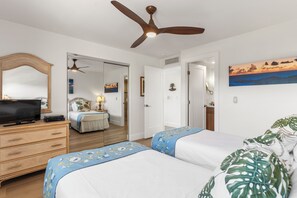 A hotel room with two beds, a television, a ceiling fan, and a painting of a sunset.