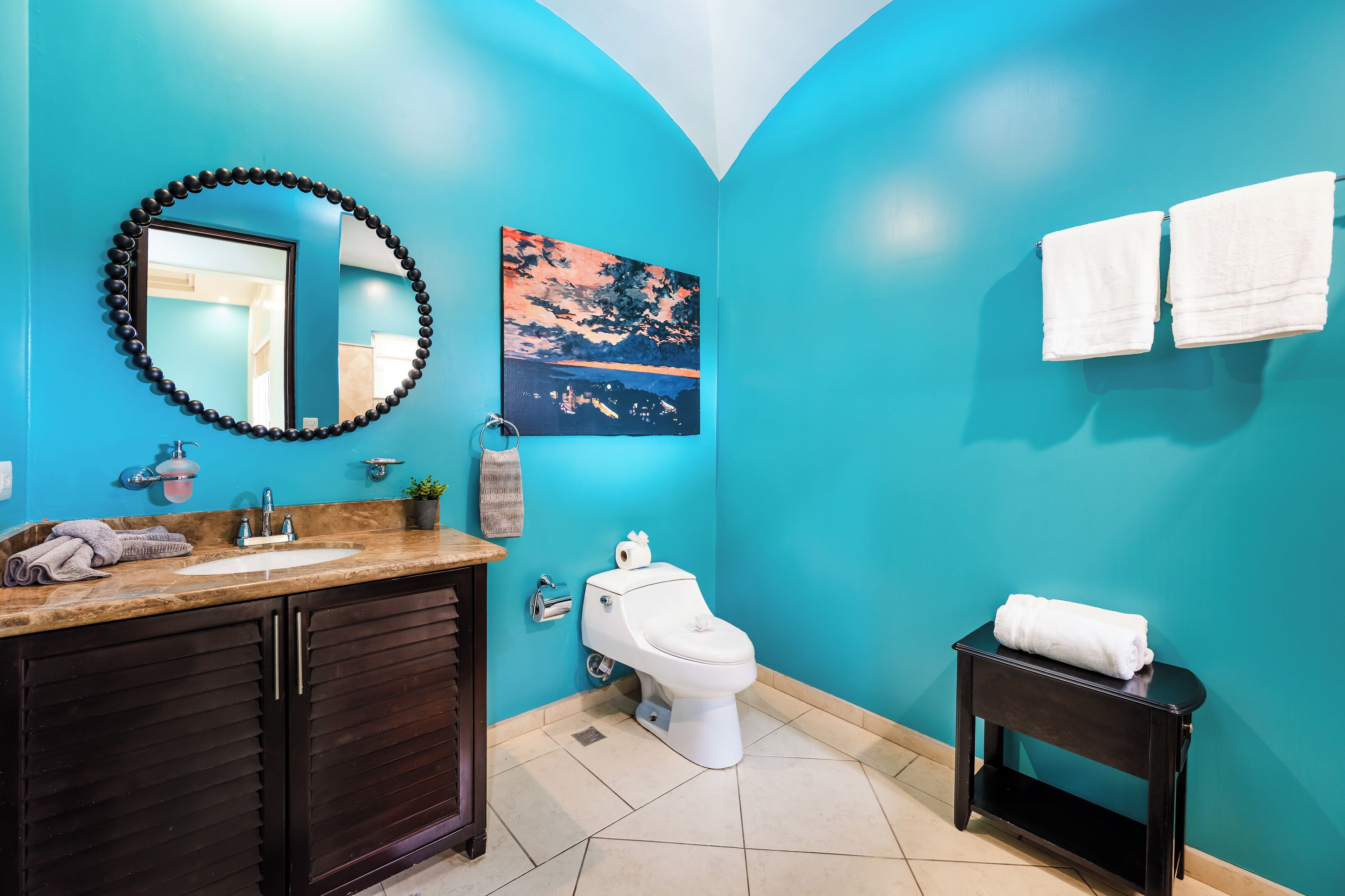 A bathroom with a blue wall, a sink with a mirror, a toilet, and a small table with towels.