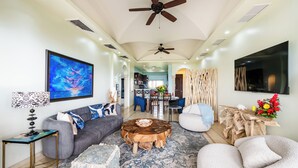 A spacious living room with a grey sofa, a round coffee table, and a white armchair. A large painting and a flat-screen TV are mounted on the walls. - Matapalo 502♆Oceanview in the 🎔 of town♆ Huge Pool Beach acss (Tamarindo)