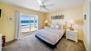 A bedroom with a bed, two bedside tables, a ceiling fan, a view of the ocean, and a small shelf with decorative items. - Just Coastin: Great Gulf Views, Screened Porch, Pet Friendly (Cape San Blas)