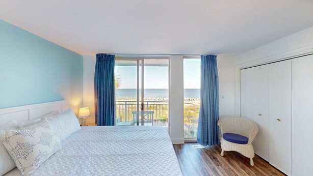 Un dormitorio con cama, mesita de noche, lámpara, silla y vista a la playa a través de una puerta corredera.