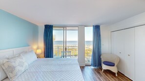 A bedroom with a bed, a nightstand, a lamp, a chair, and a view of the beach through a sliding door.