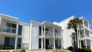 Un hotel blanco de varios pisos con balcones, mobiliario exterior y una palmera.