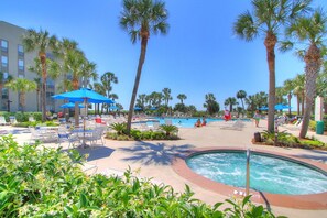 A resort pool area with lounge chairs and umbrellas, surrounded by palm trees.