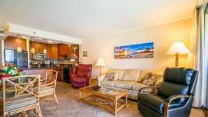 A living room with a sofa, two chairs, a coffee table, and a kitchen area with wooden cabinets and appliances.