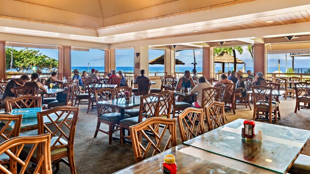 A spacious outdoor dining area with wooden chairs and tables, large windows offering views of the ocean, and a ceiling fan.