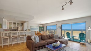 A spacious living area with a brown sofa, dining chairs, a coffee table, and a view of the beach through large windows. - 1213O Local's Choice at Silverstrand (Oxnard)