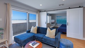A room with a blue sofa, a small ottoman, a remote control on a table, and a view of the sea through large windows. - 1213O Local's Choice at Silverstrand (Oxnard)