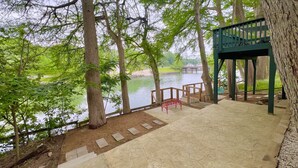 A stone deck with a red bench overlooking a lake, surrounded by trees. - Riverside Roost- NB Guadalupe Riverfront + Easy River Access + Upper & Lower Decks + Minutes to Town (New Braunfels)
