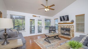 A spacious living room with a fireplace, a ceiling fan, and a large window overlooking a deck. - Riverside Roost- NB Guadalupe Riverfront + Easy River Access + Upper & Lower Decks + Minutes to Town (New Braunfels)