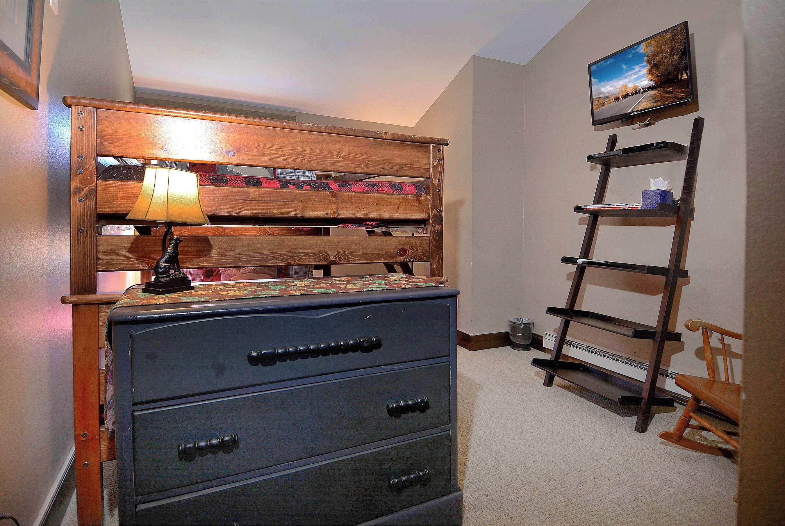A bedroom with a wooden headboard, a dresser, a TV mounted on the wall, and a ladder shelf.