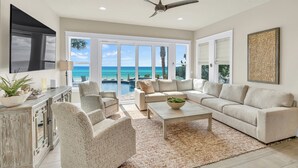 A spacious living room with a large sofa, a coffee table, and a TV. There is a ceiling fan and a view of the ocean through large windows. - Not the Norm - Oceanfront Home with Private Pool & Beach Access (Miramar Beach)