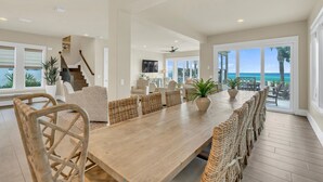A spacious dining area with a long wooden table and wicker chairs, a staircase, and a view of the ocean. - Not the Norm - Oceanfront Home with Private Pool & Beach Access (Miramar Beach)