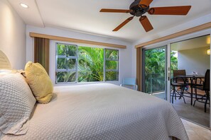 Ein Hotelzimmer mit Bett, Deckenventilator, Balkon mit Tisch und Stühlen sowie großen Fenstern mit Blick auf Palmen.