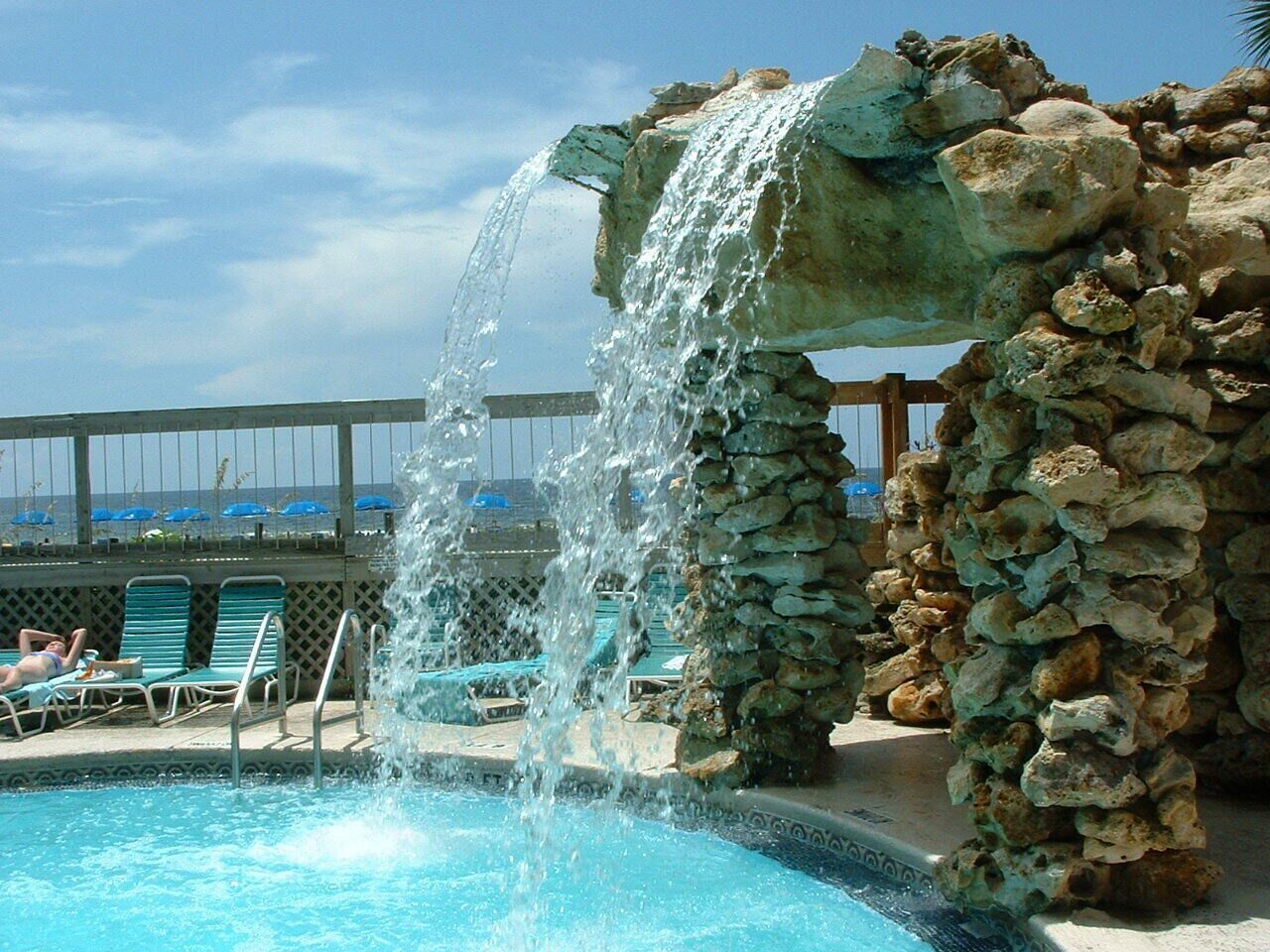 A pool with a waterfall feature, surrounded by lounge chairs and a stone wall.